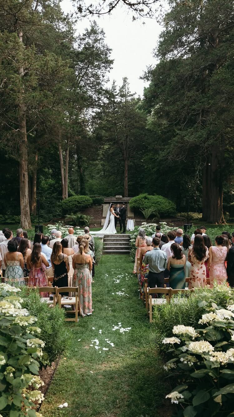 Outdoor wedding ceremony in a forest with guests seated along a flower-lined aisle.