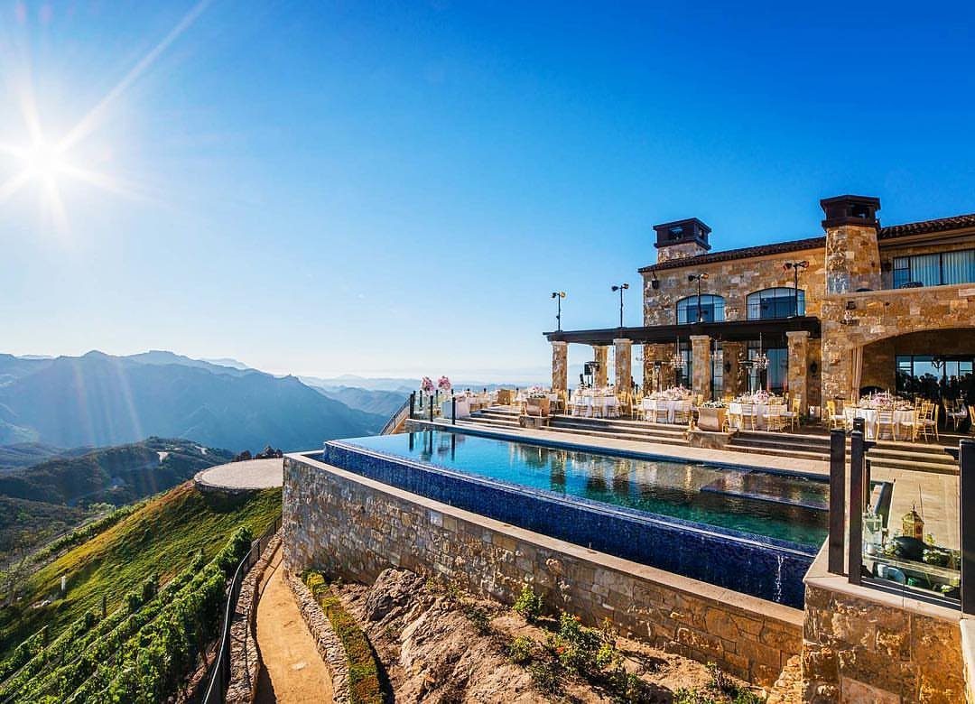 Stone mansion with infinity pool overlooking mountains and vineyards under a sunny sky