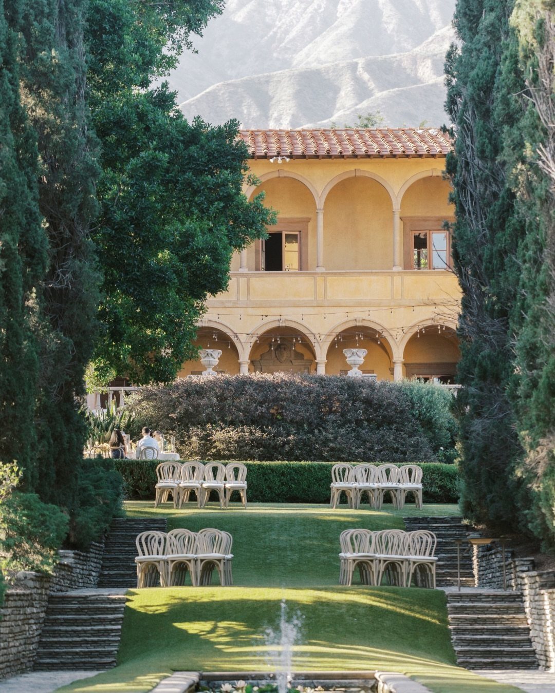 Yellow Mediterranean villa with white ceremony chairs on a green lawn and mountains.