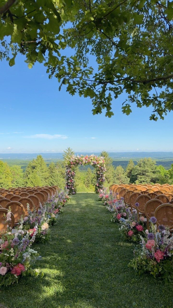Outdoor wedding ceremony setup with wooden chairs and floral arch overlooking green hills.