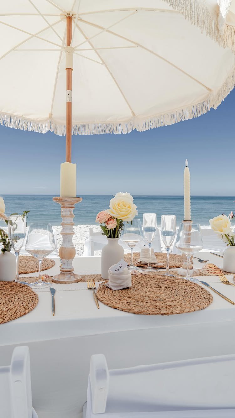 Elegant beach table setting with white roses and umbrella overlooking the blue ocean.