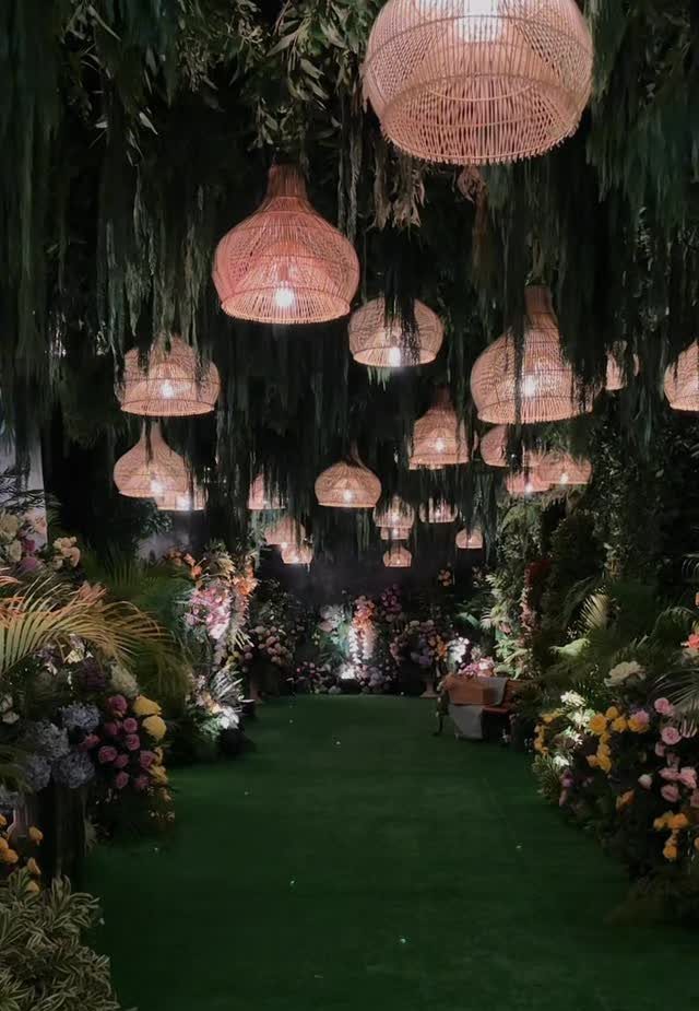 Floral aisle with green carpet and woven wicker lamps under hanging foliage
