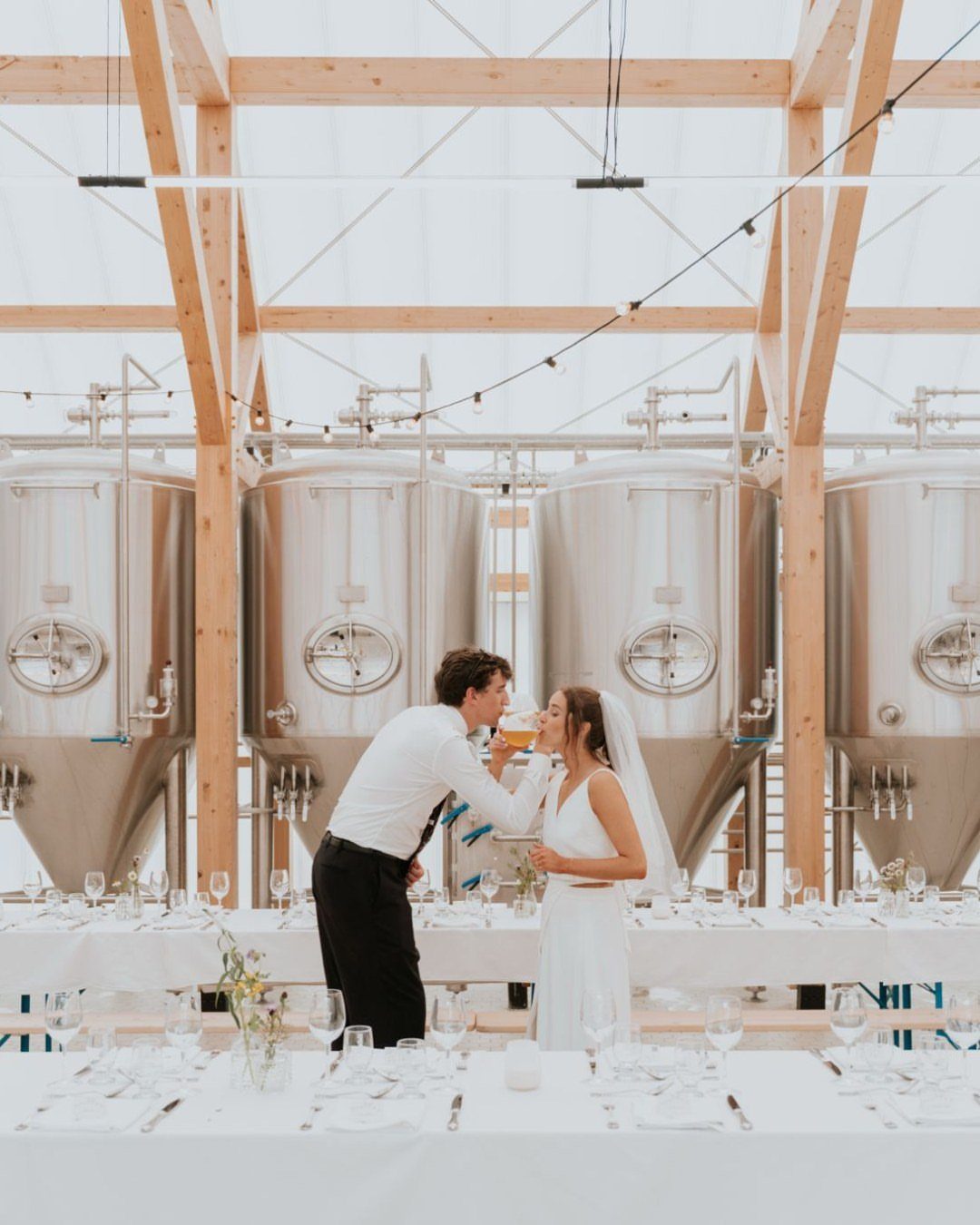 Bride and groom sharing a drink in a brewery with large steel tanks.