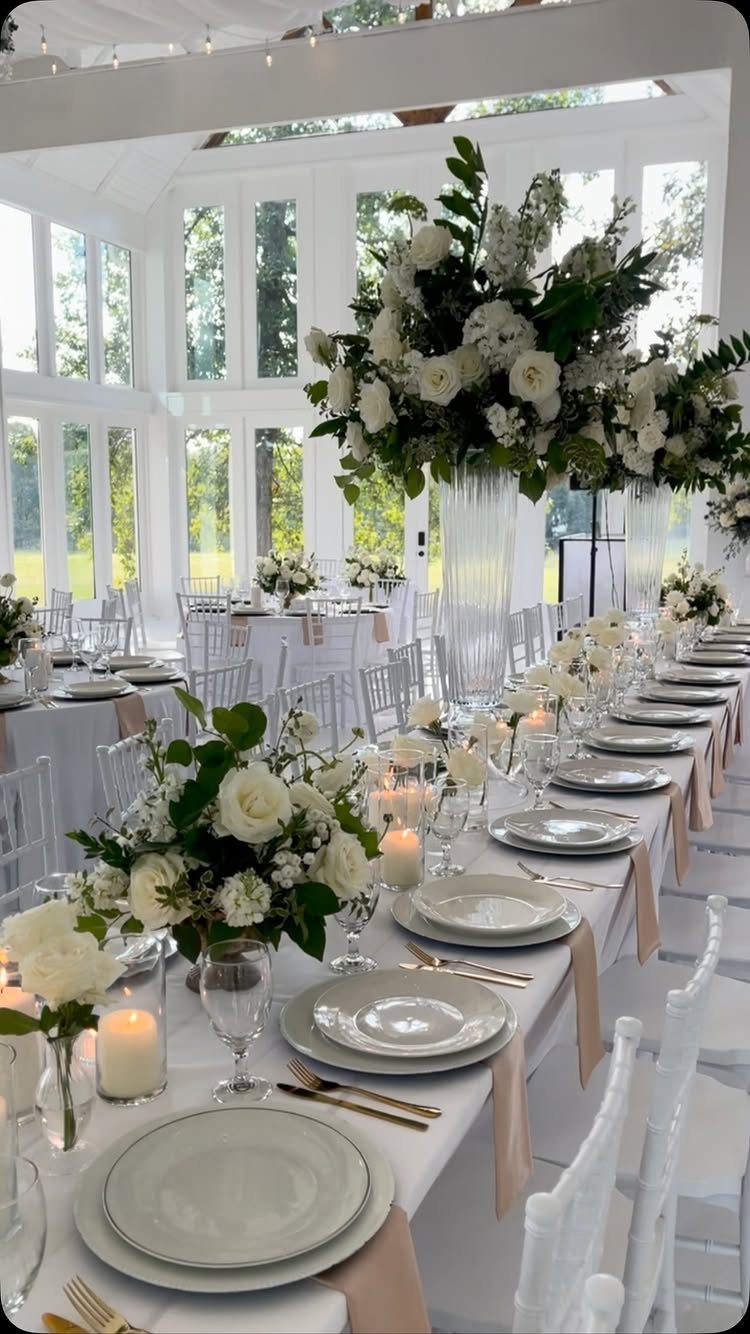 Long white reception table with floral centerpieces inside a bright white glasshouse venue