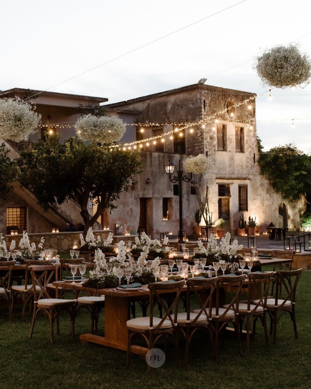 Outdoor reception tables with white flowers and string lights before a historic stone estate.