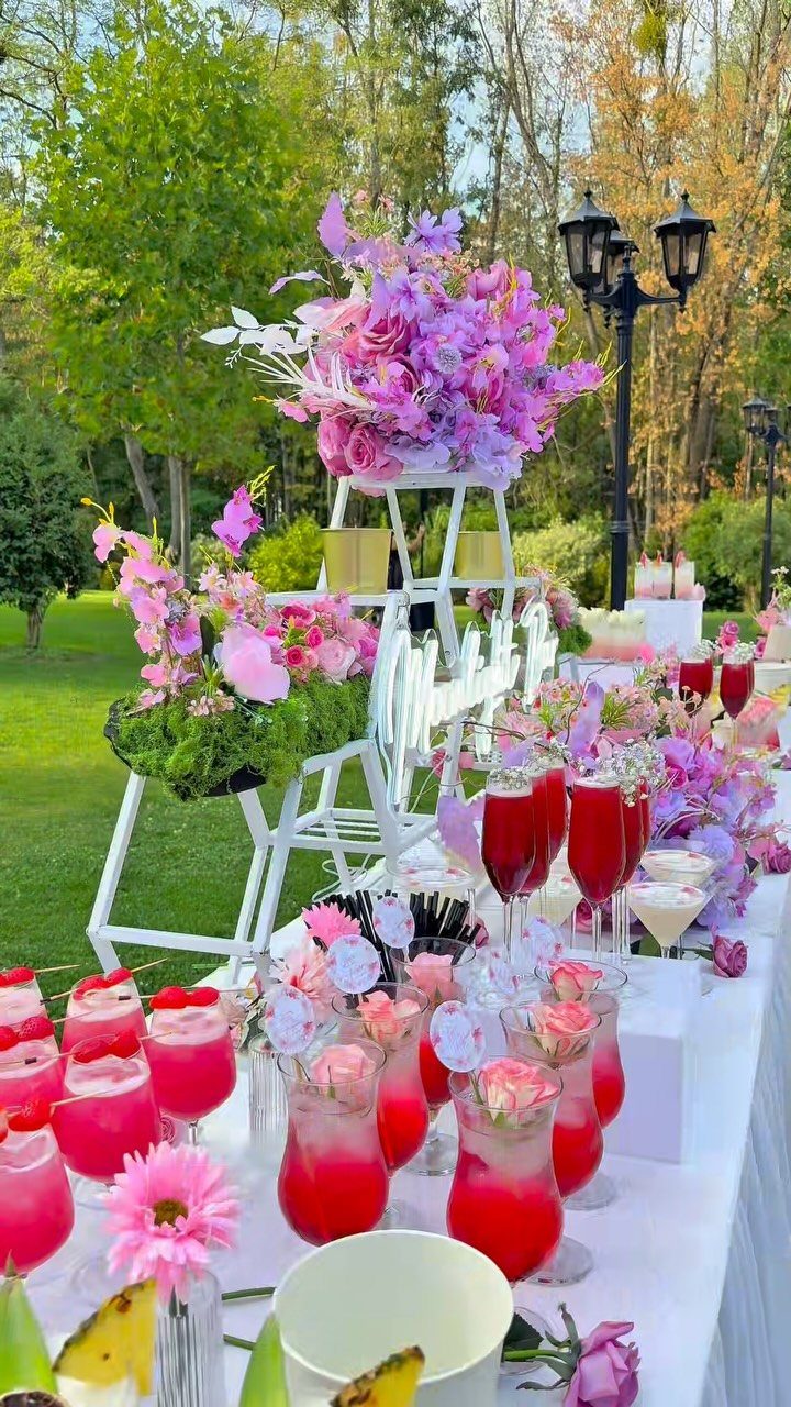 Pink drinks and floral arrangements on a white table at an outdoor reception.