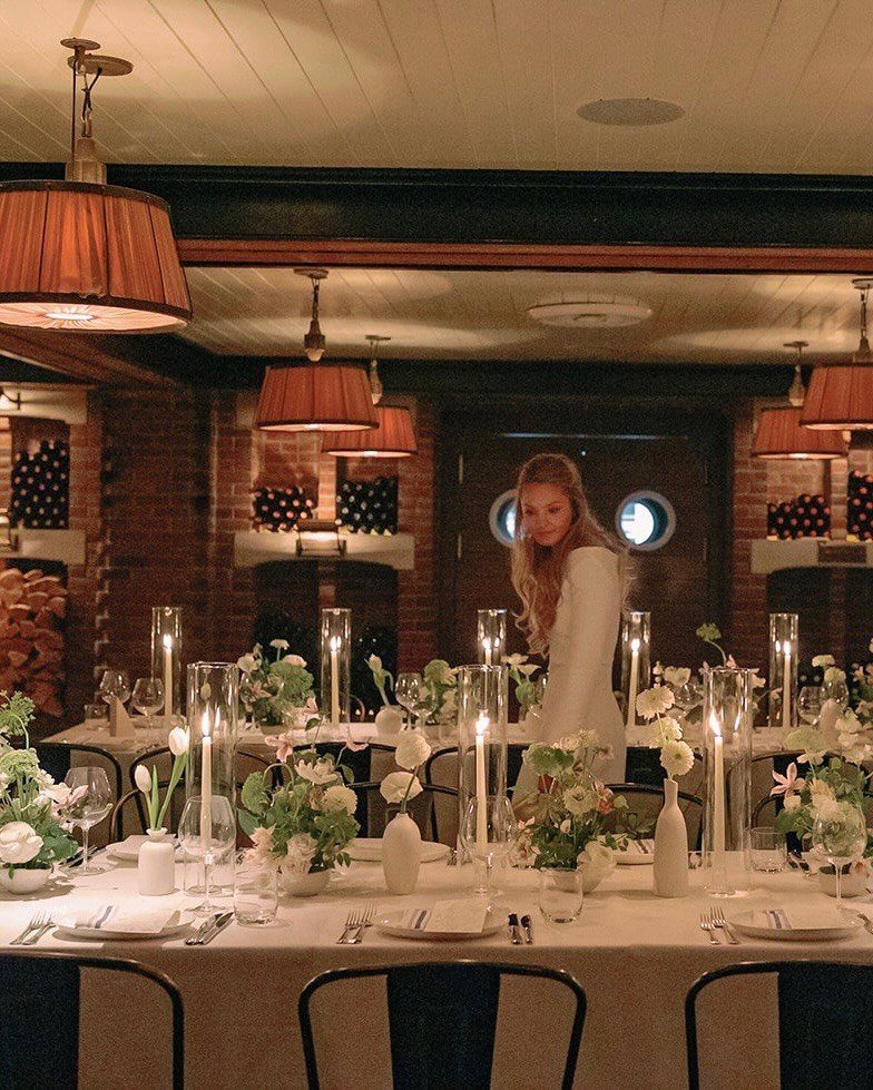 Woman in white dress standing by elegantly set dinner tables with candles and flowers.