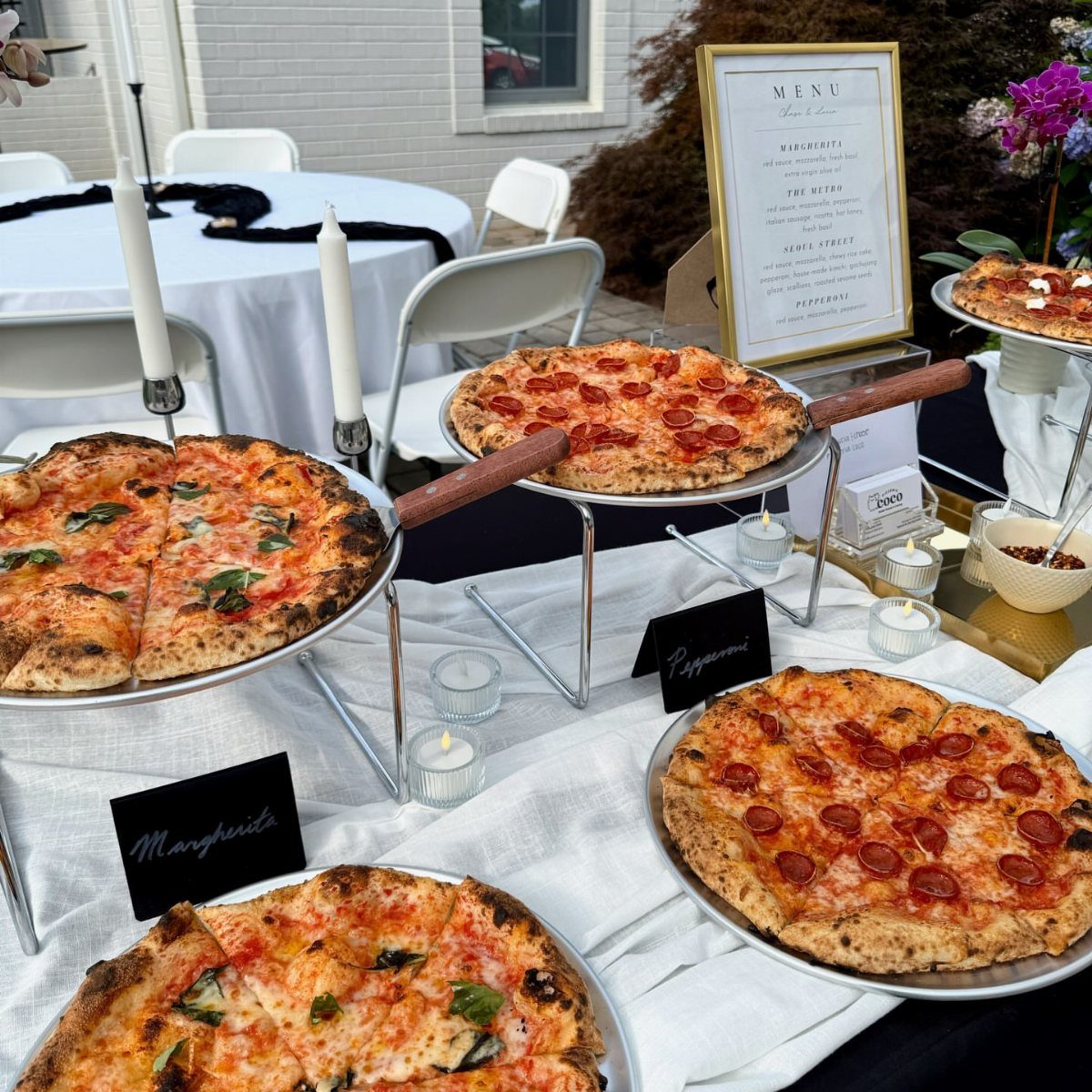 Pizzas on metal stands and a menu on a white outdoor catering table