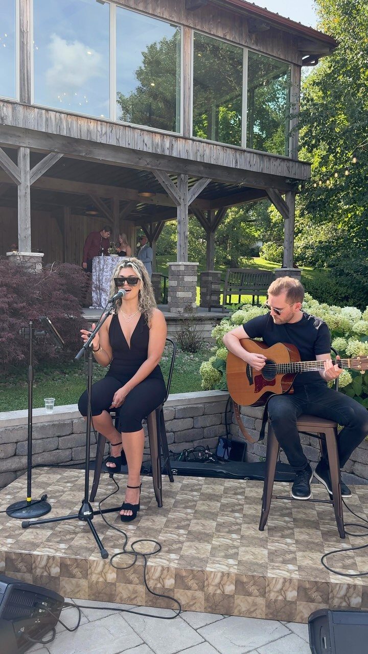 Musicians performing live music on an outdoor stage with a rustic building background.