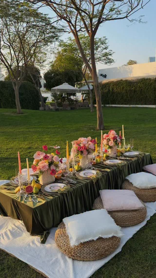 Outdoor dining table with green cloth, pink flowers, and woven pouf seating.
