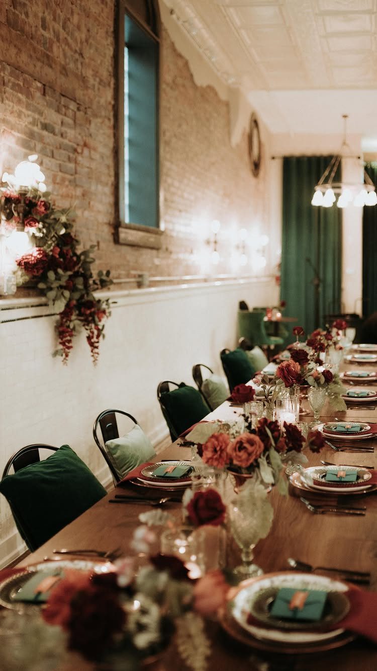 Long wooden dining table with red floral centerpieces and green cushions in brick room