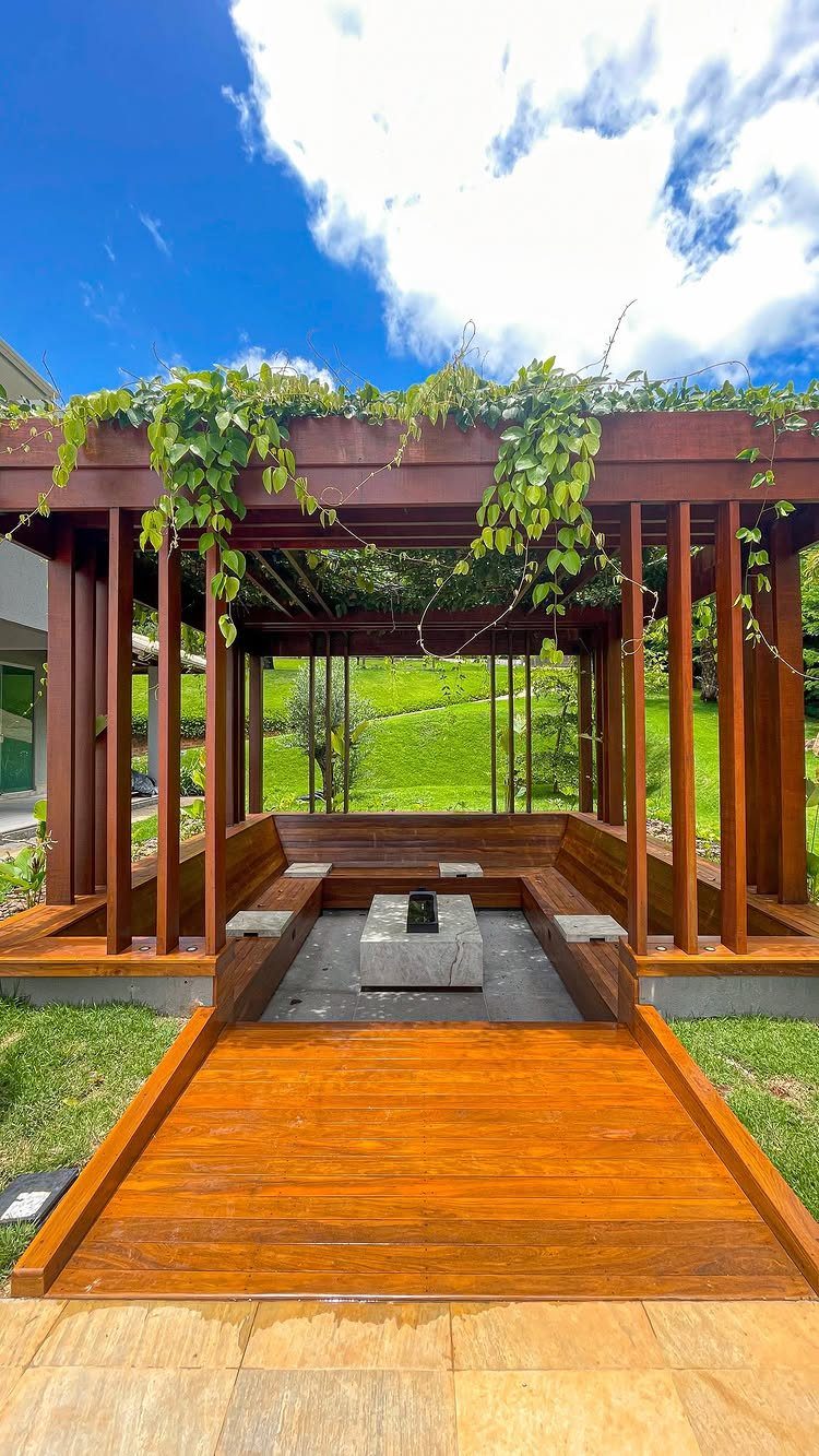 Wooden pergola with built-in seating and a stone fire pit in a backyard.
