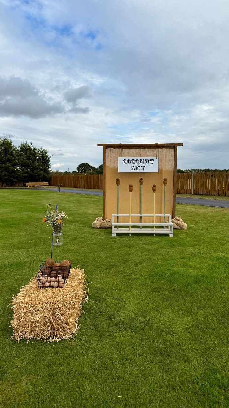 Coconut shy game setup with wooden booth and hay bale on green grass.