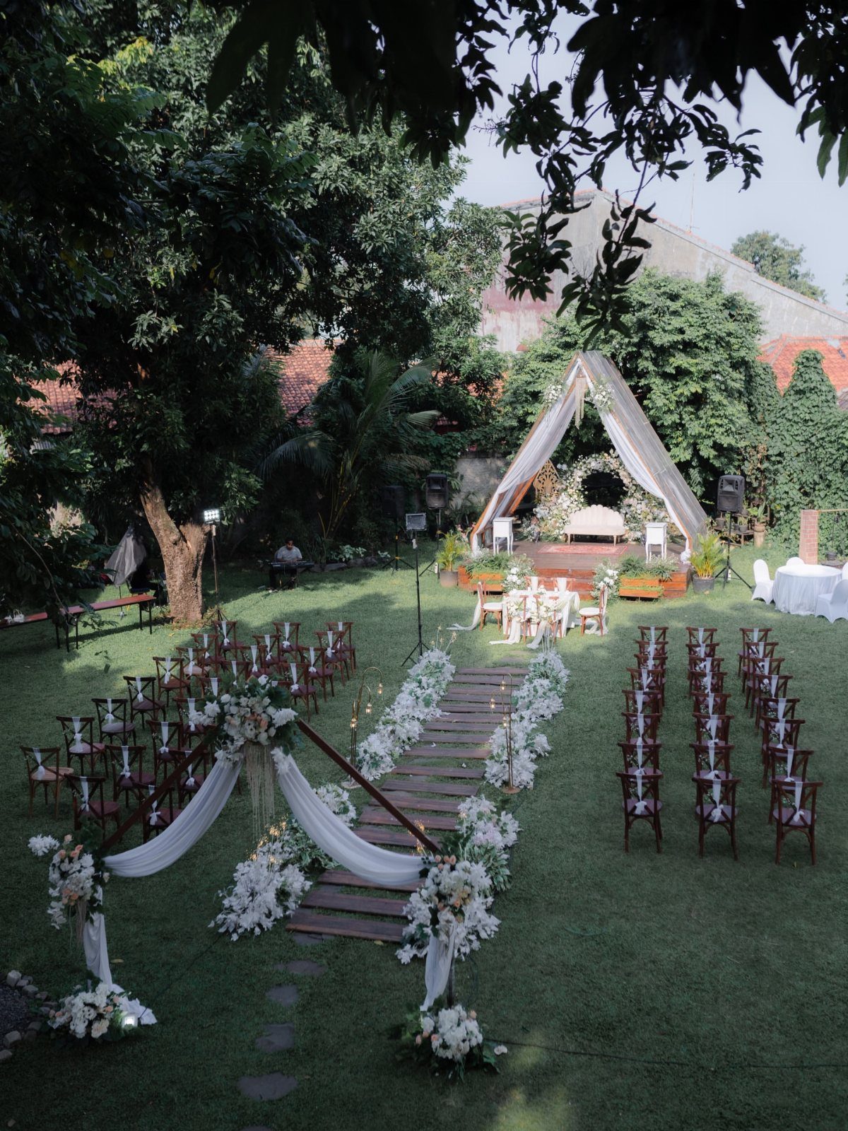 Wedding ceremony setup with a white floral arch and wooden aisle on green grass.
