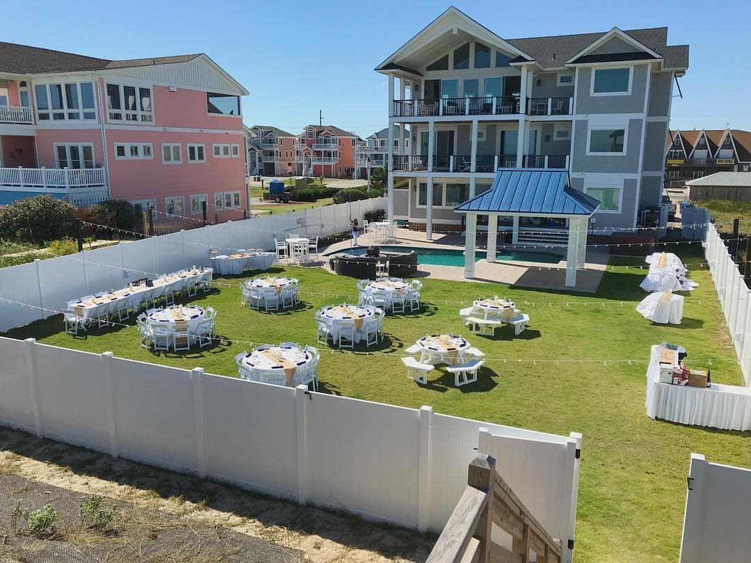 Fenced backyard event setup with white tables and yellow runners near a house.
