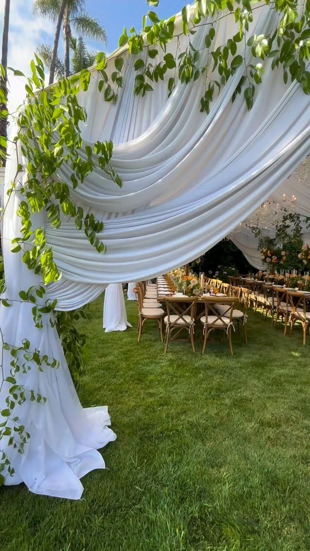 White draped canopy with green vines over wooden wedding tables on green grass.