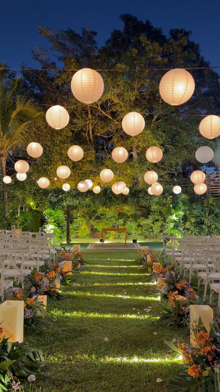 Outdoor wedding ceremony aisle with white chairs, floral arrangements, and hanging paper lanterns.