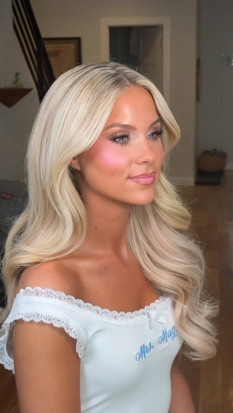 Woman with wavy blonde hair and bridal makeup wearing a white lace top.