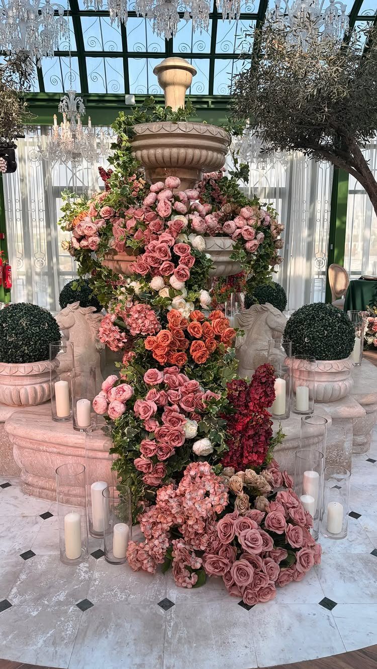 Tiered stone fountain covered in pink roses inside a glass greenhouse with chandeliers.