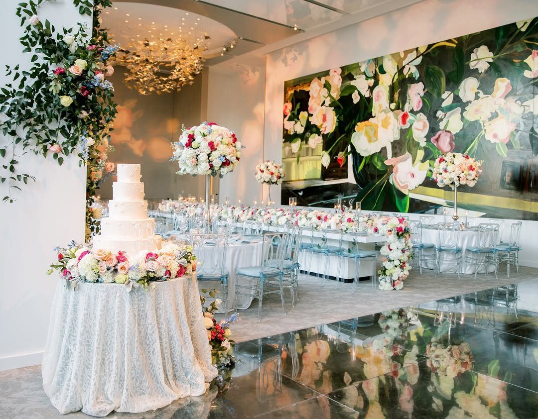 Wedding reception setup with a tiered cake, floral mural, and clear ghost chairs.