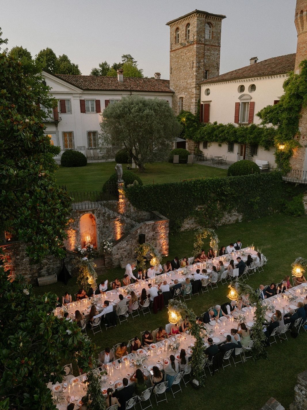 Long dinner tables set in a historic courtyard with stone buildings and tower.