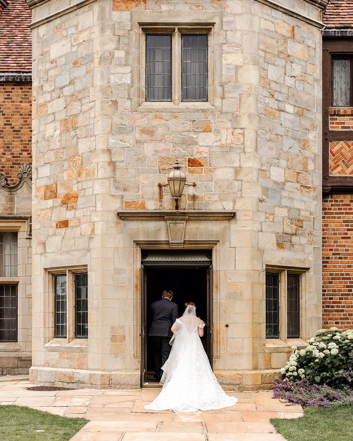 Bride and groom entering a grand stone mansion with a turret and windows.