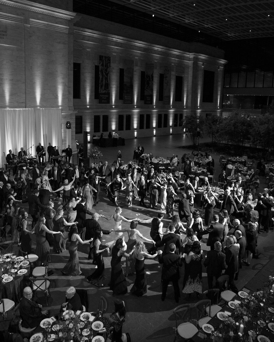 Formal gala reception with guests dancing in a grand stone atrium with high ceilings.