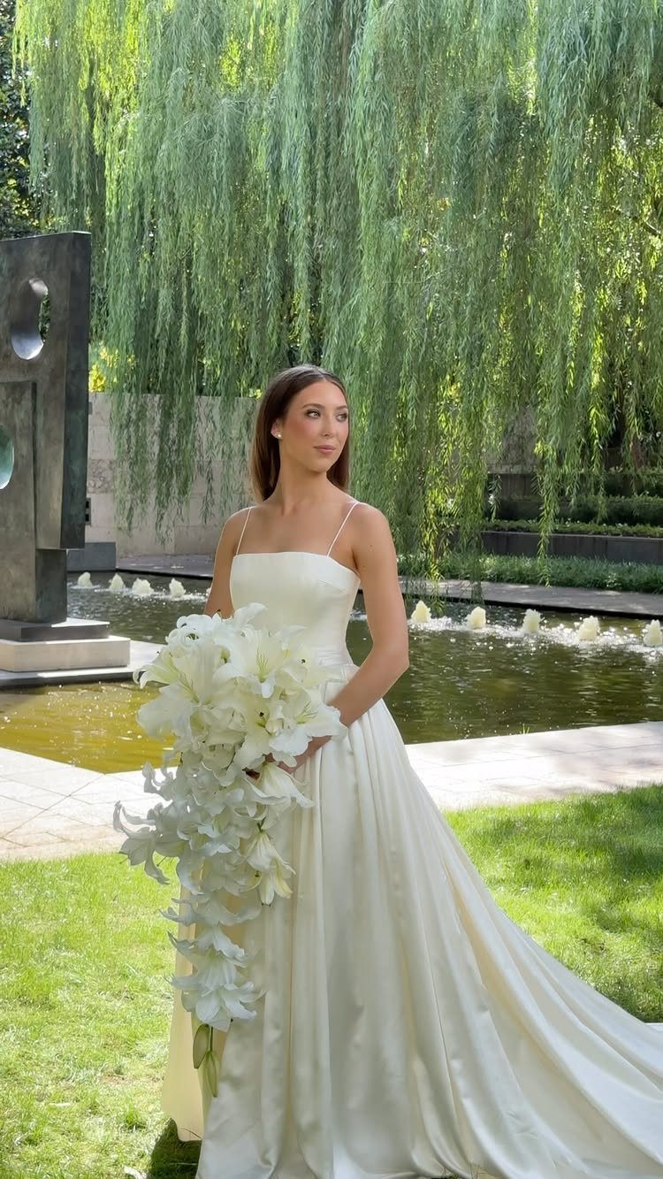Bride in white gown holding orchids before a sculpture and weeping willow tree
