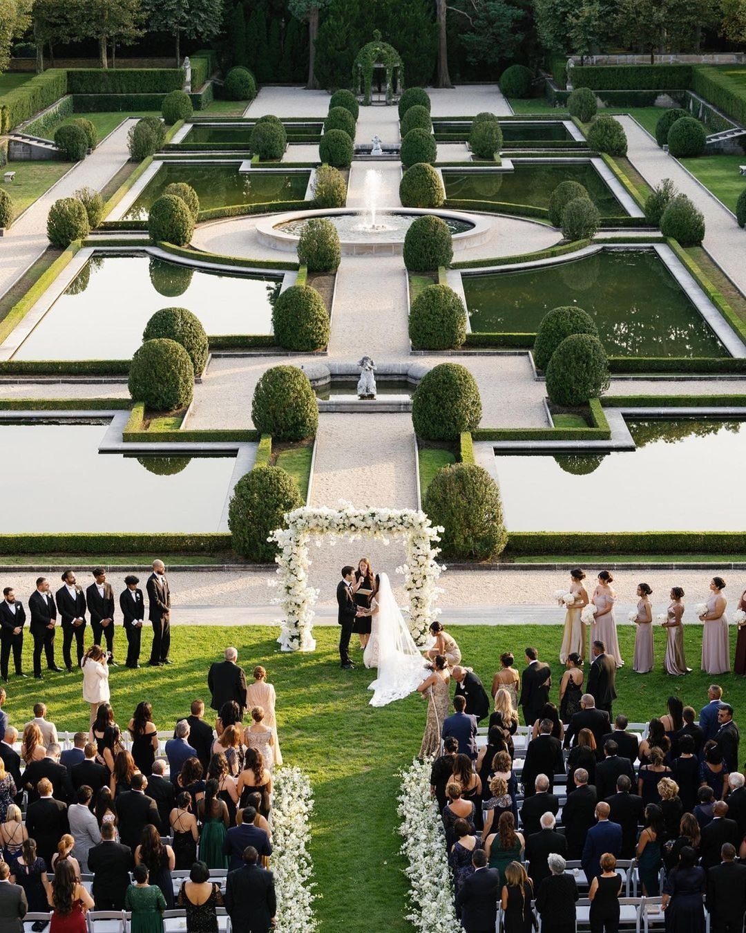 Wedding ceremony in a formal French garden with symmetrical ponds and manicured hedges.