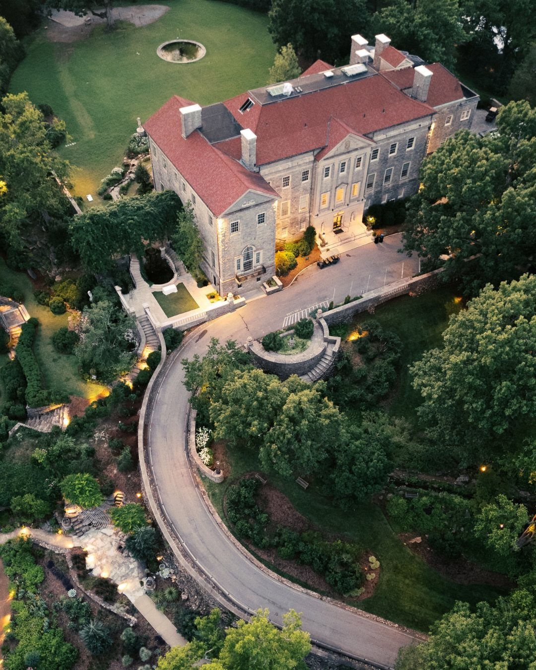 Large stone mansion with red roof surrounded by lush green gardens and driveway