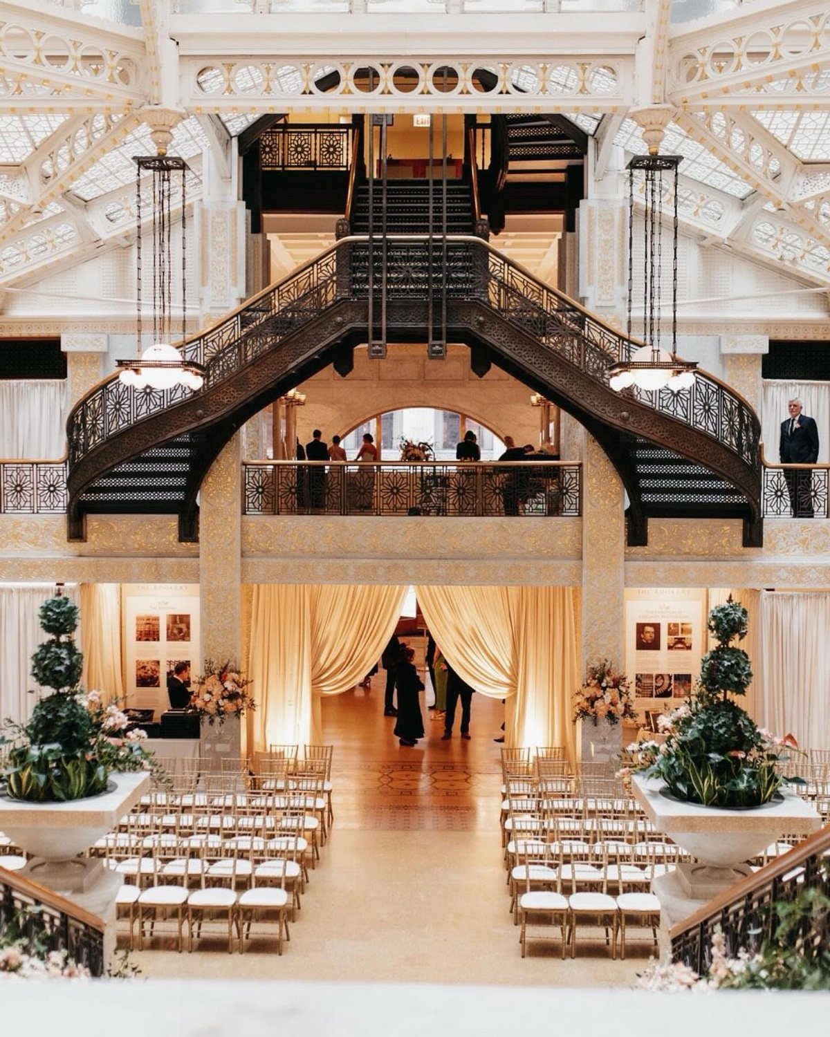 Grand ornate metal staircase overlooking a wedding ceremony setup with gold chairs.