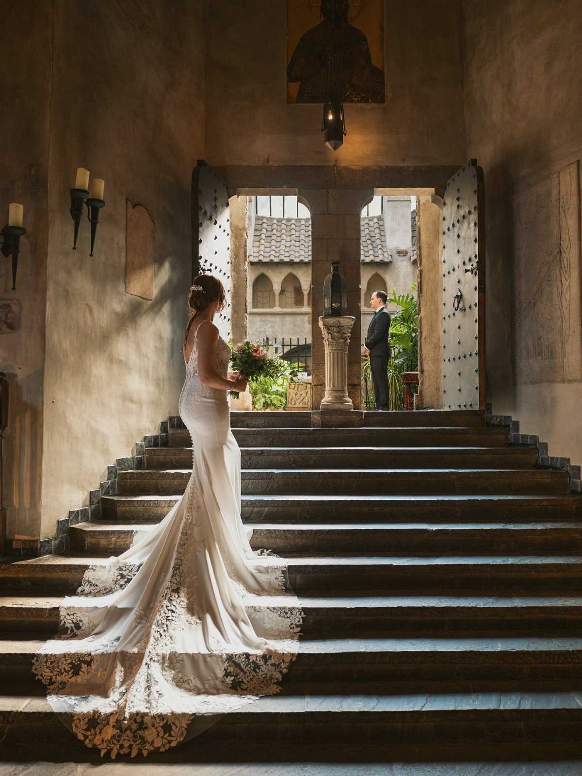 Bride in white gown on stone stairs facing groom in castle archway
