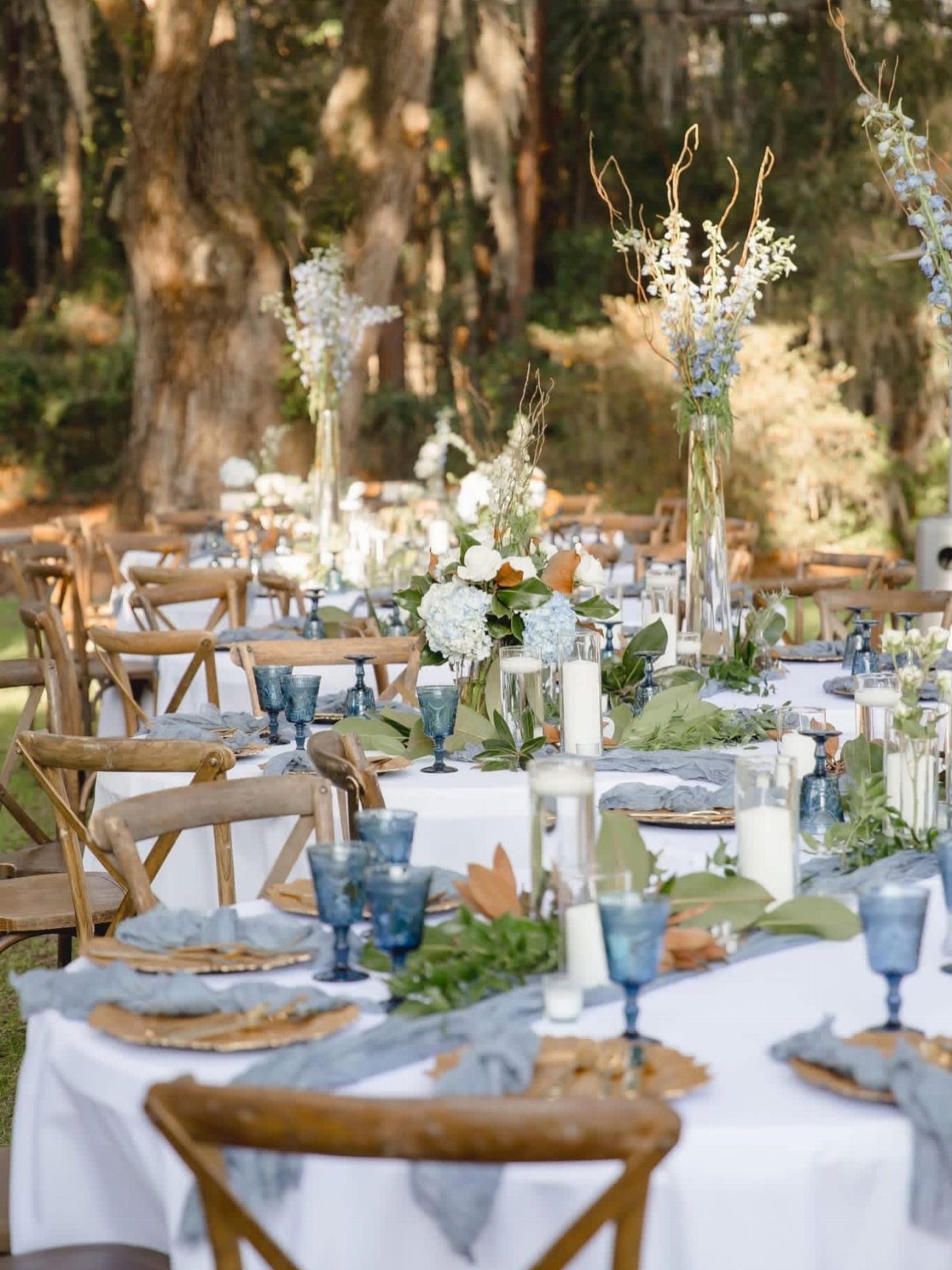 Outdoor wedding reception tables with blue accents set under large mossy oak trees.