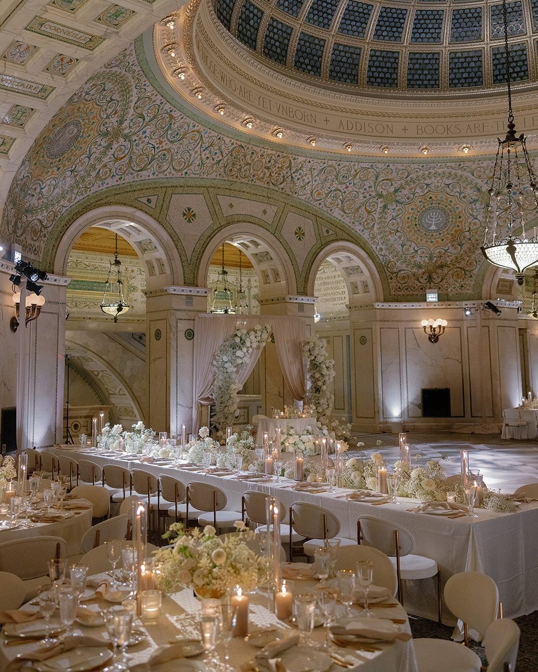 Ornate domed venue with long white tables, floral centerpieces, and glowing candles.