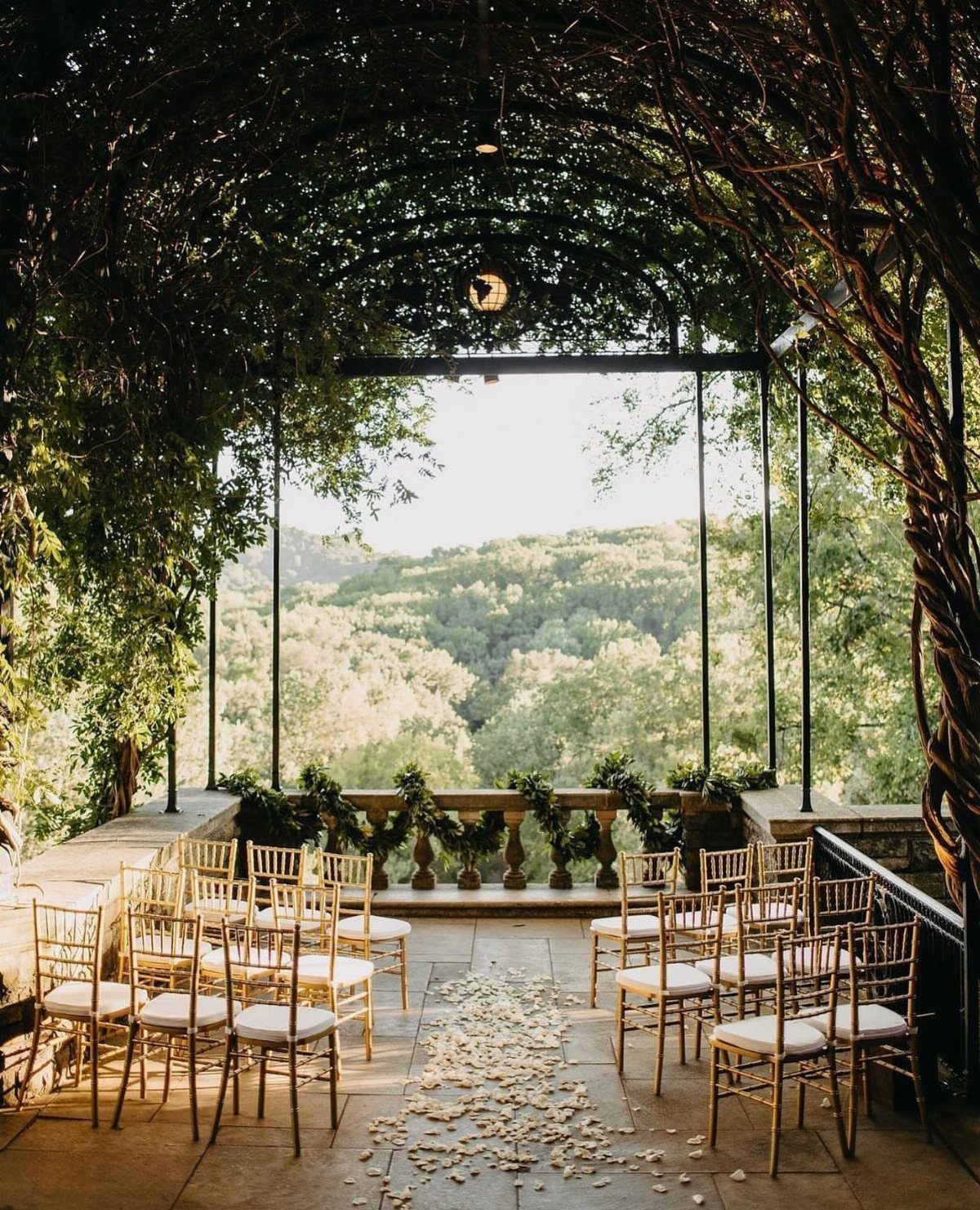 Outdoor wedding ceremony setup with gold chairs under a vine trellis overlooking hills