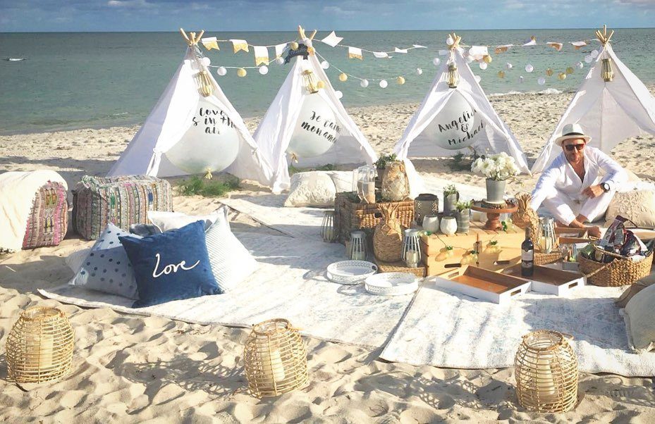 Beach picnic setup with white teepees, rugs, and a man in white clothing.