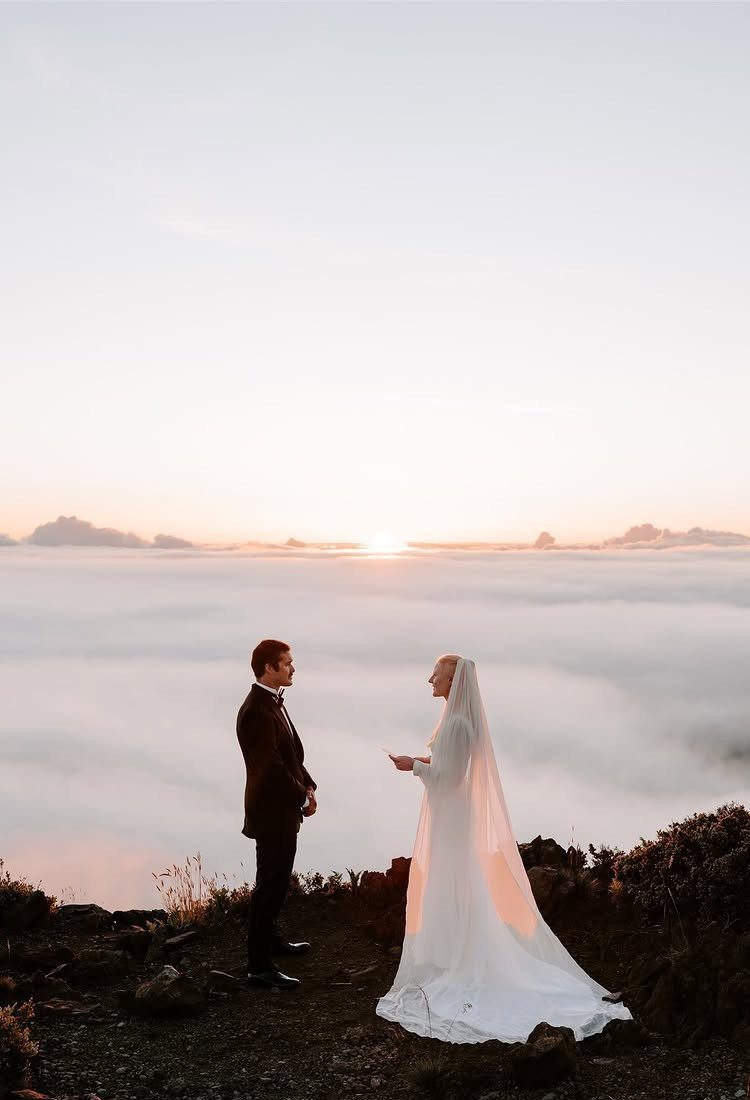 Couple in wedding attire facing each other on a mountain peak at sunrise.