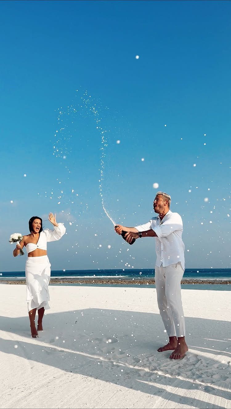 Couple in white celebrating on a beach with a champagne bottle spray.
