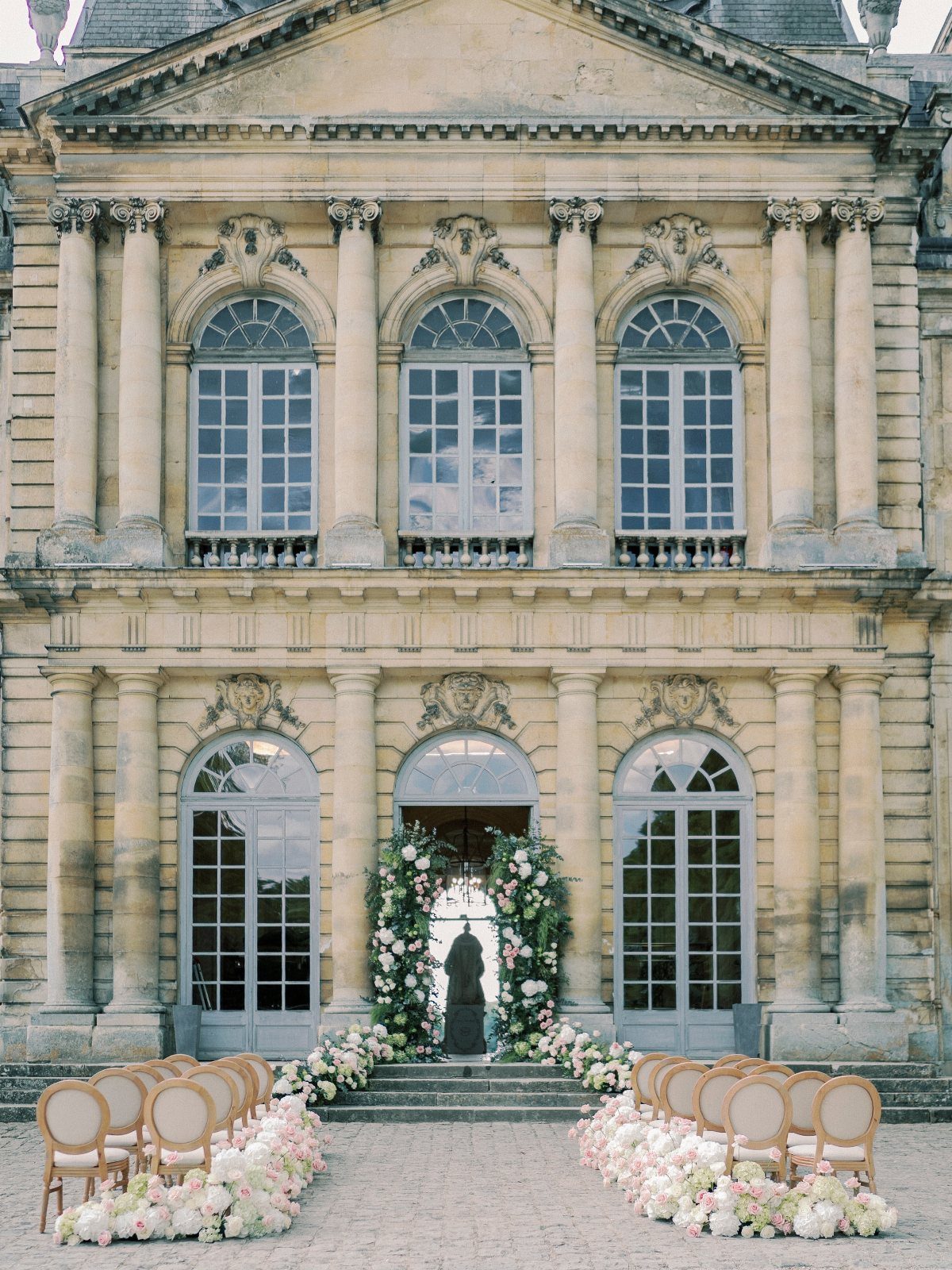 Outdoor ceremony setup with beige chairs and floral arrangements before a classical stone building.