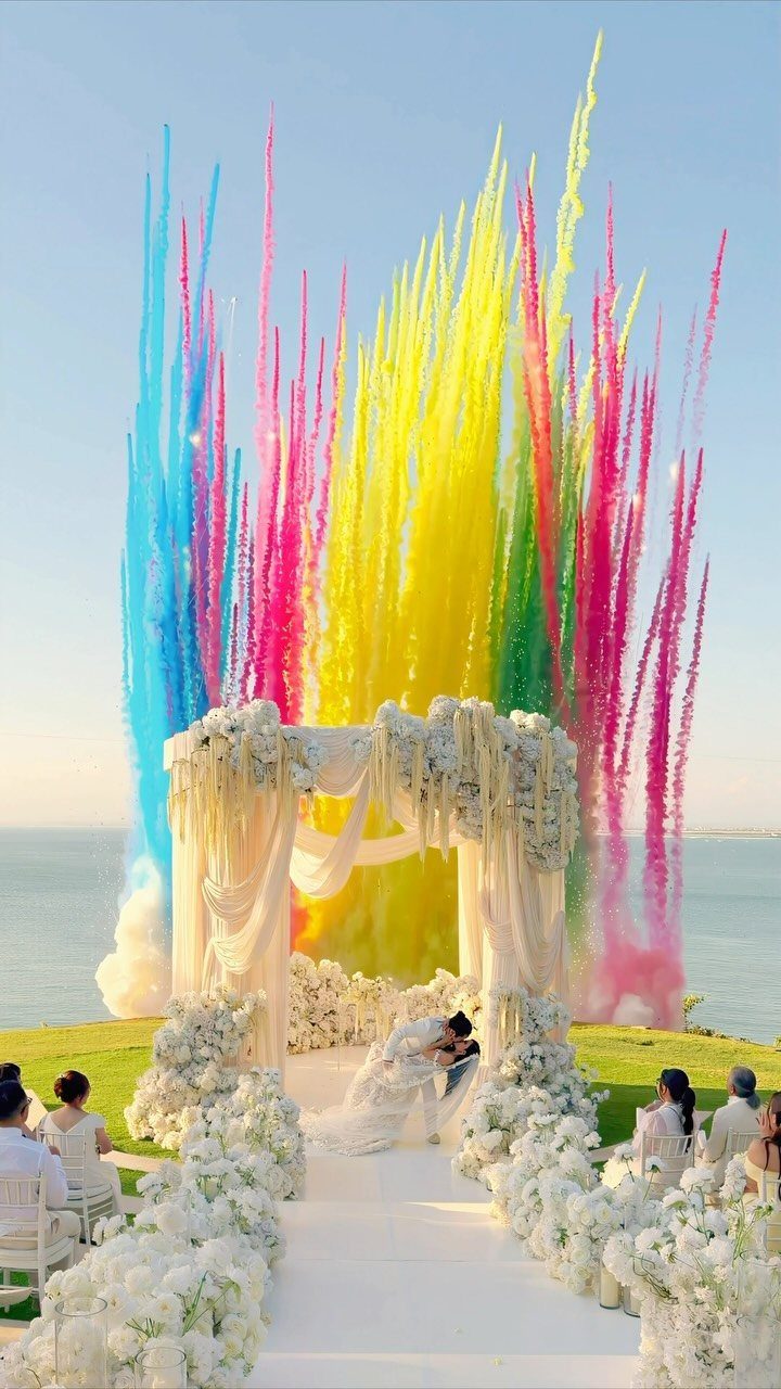 Couple kissing under a white floral arch with rainbow smoke plumes behind them.