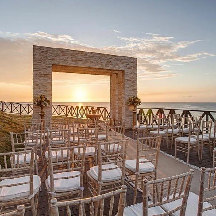 Outdoor ceremony setup with stone arch and wooden chairs overlooking ocean at sunset