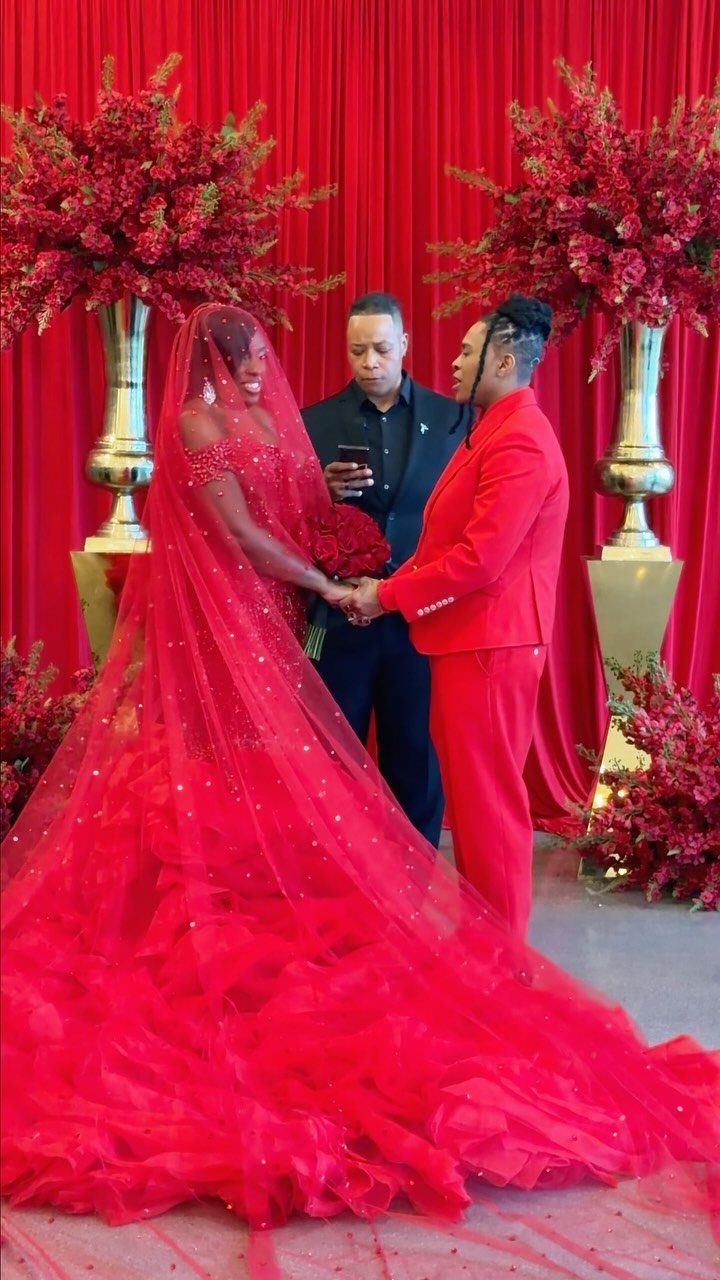 Couple in red formal wear holding hands during a ceremony with red floral decor.