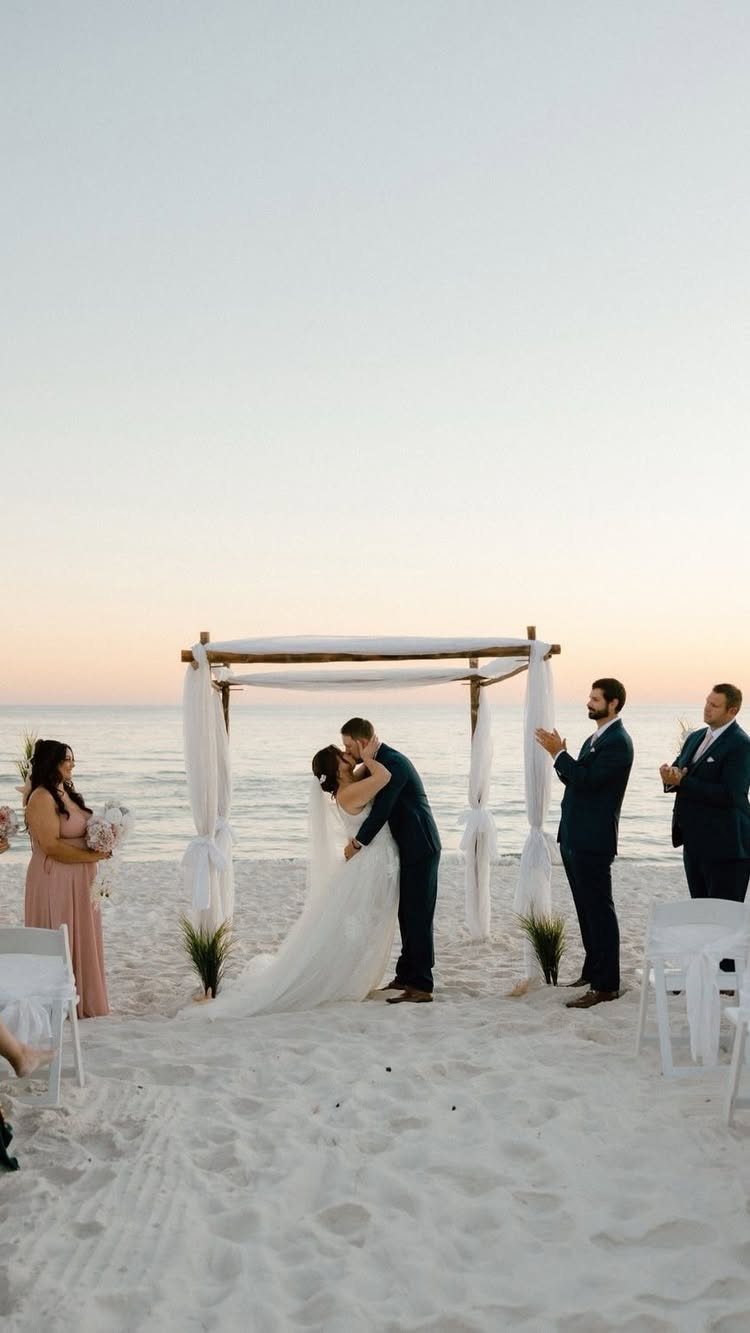 Bride and groom kissing under a white canopy on a sandy beach at sunset.