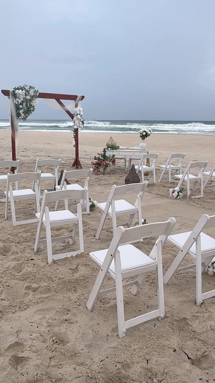 White folding chairs and a floral wooden arch on a sandy beach shoreline.