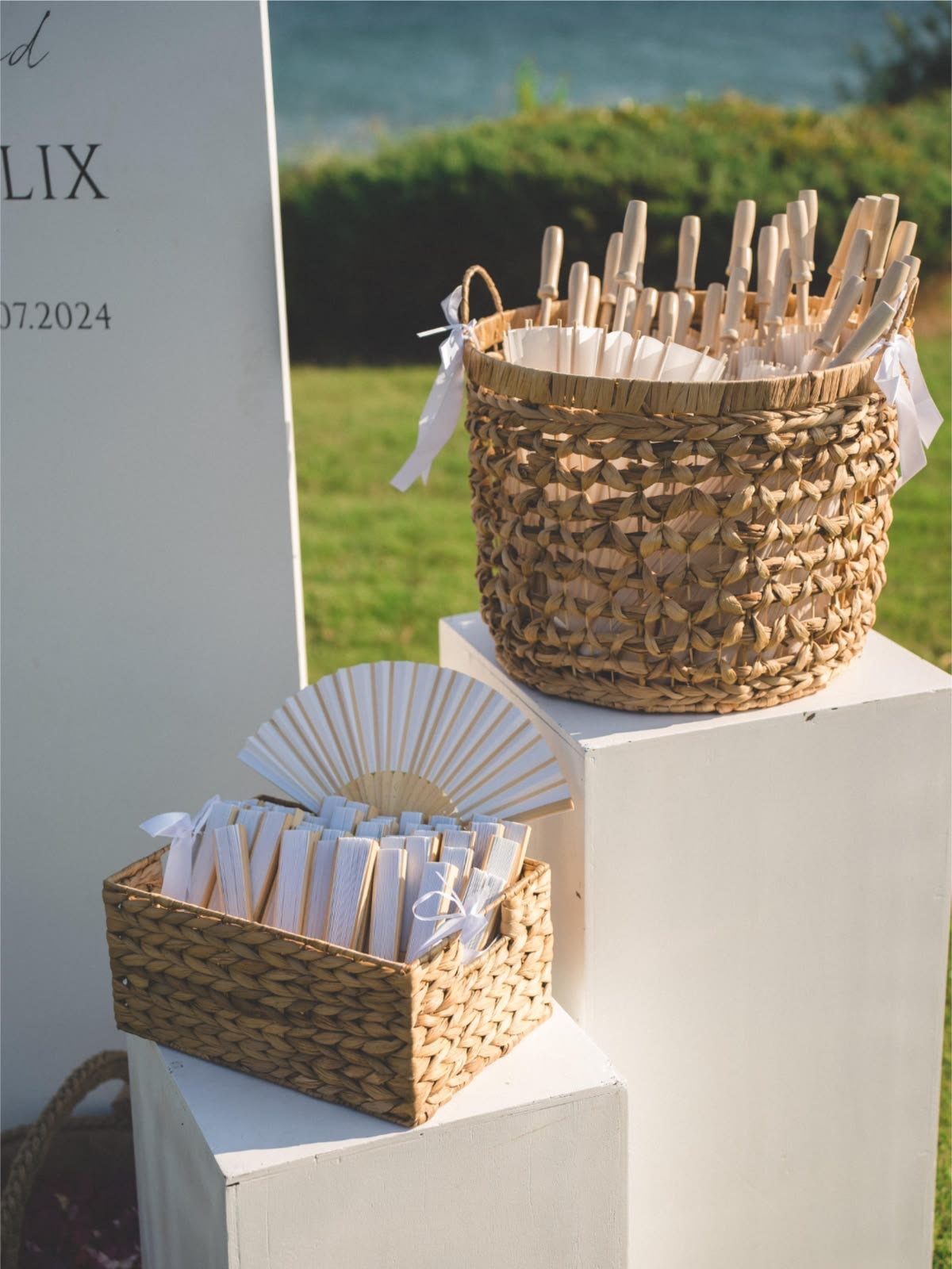 Woven baskets with white fans and programs on pedestals at a beach wedding.