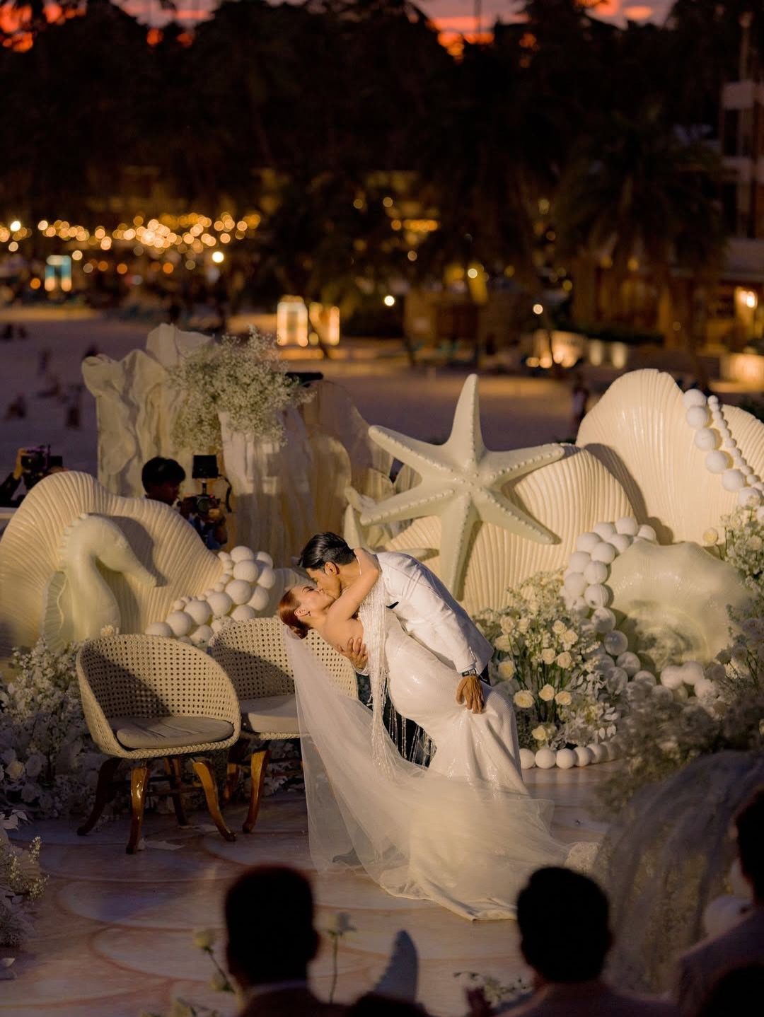 Bride and groom kissing at a beach wedding ceremony with giant seashell decorations.