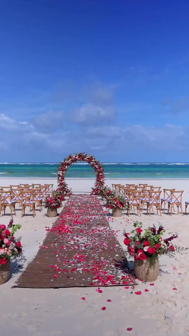 Beach wedding ceremony setup with floral arch, red petal aisle, and wooden chairs.