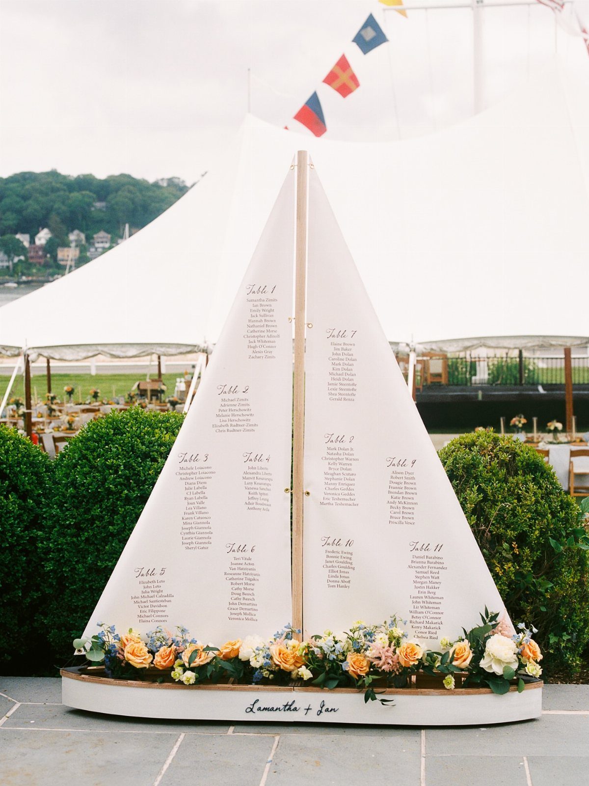 Sailboat-shaped wedding seating chart decorated with flowers on a stone patio outdoors.