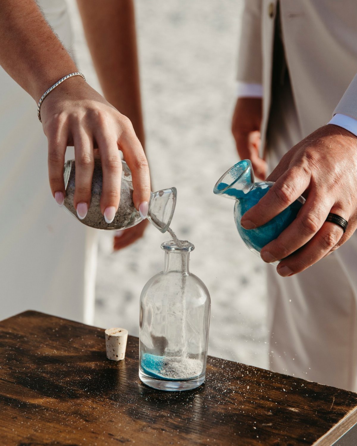 Hands pouring white and blue sand into a glass bottle on a beach.