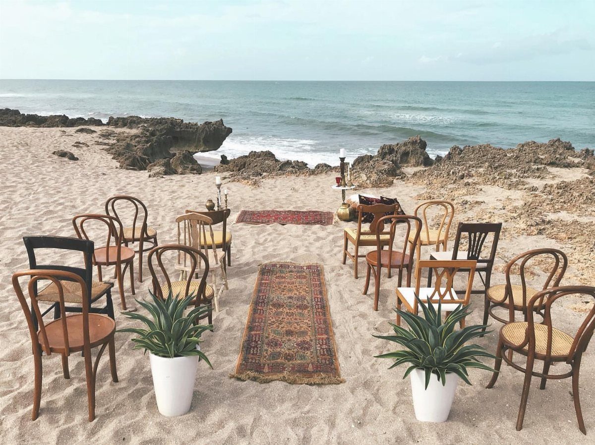 Mismatched wooden chairs and patterned rugs arranged for a wedding ceremony on a beach.